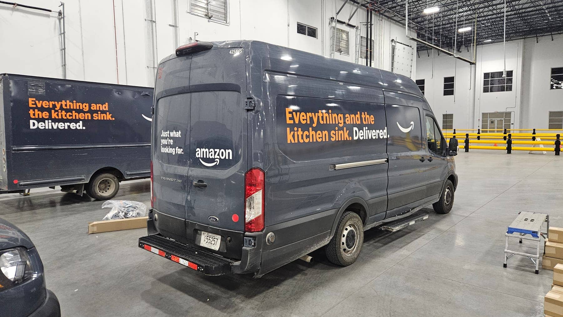 Dark gray Ford Transit with Amazon smile logo and kitchen-sink campaign graphics in a warehouse