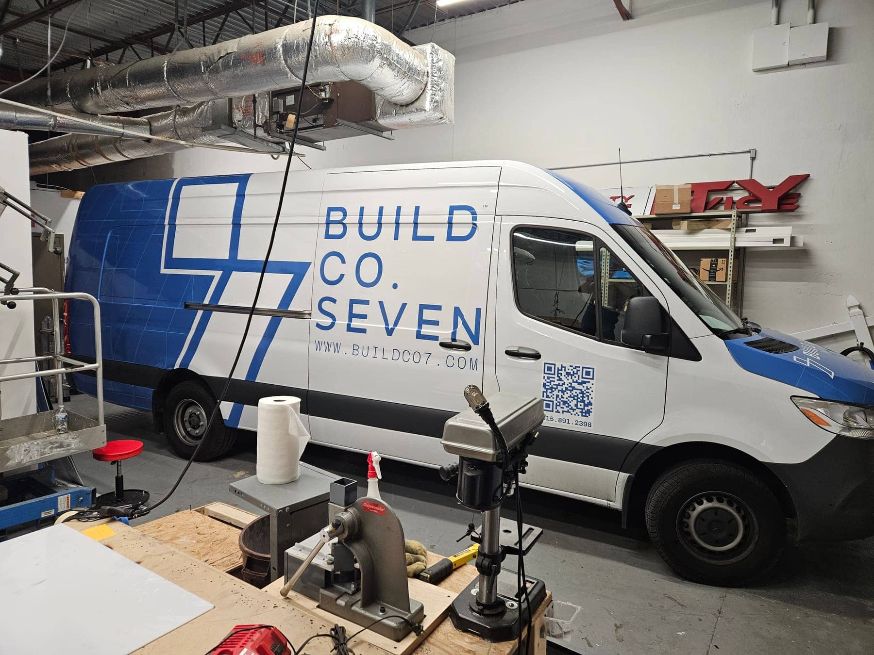 White Mercedes Sprinter with large blue Build Co Seven graphics and QR code, in the install bay