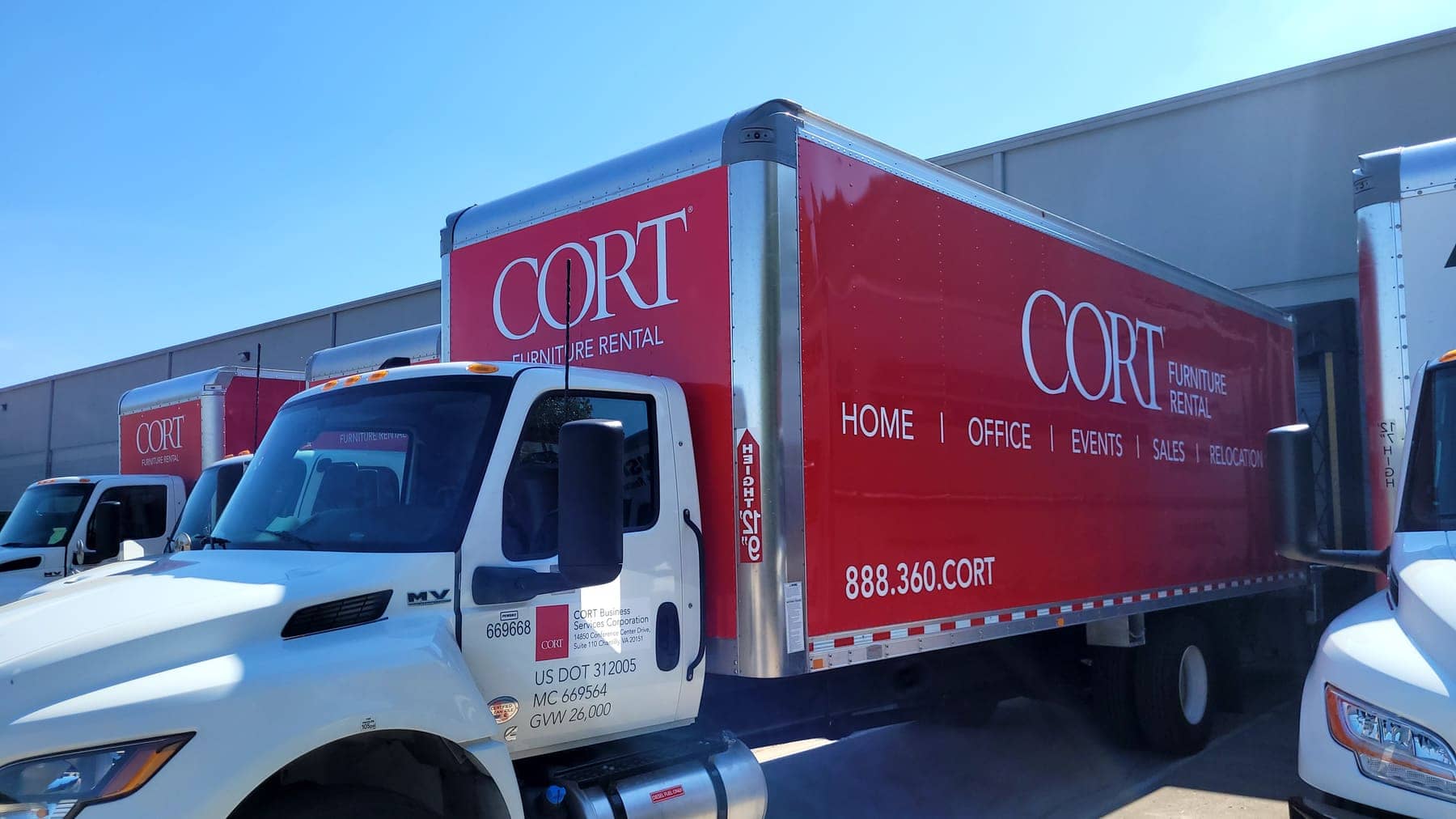 Two red CORT Furniture Rental box trucks with white lettering at a loading area
