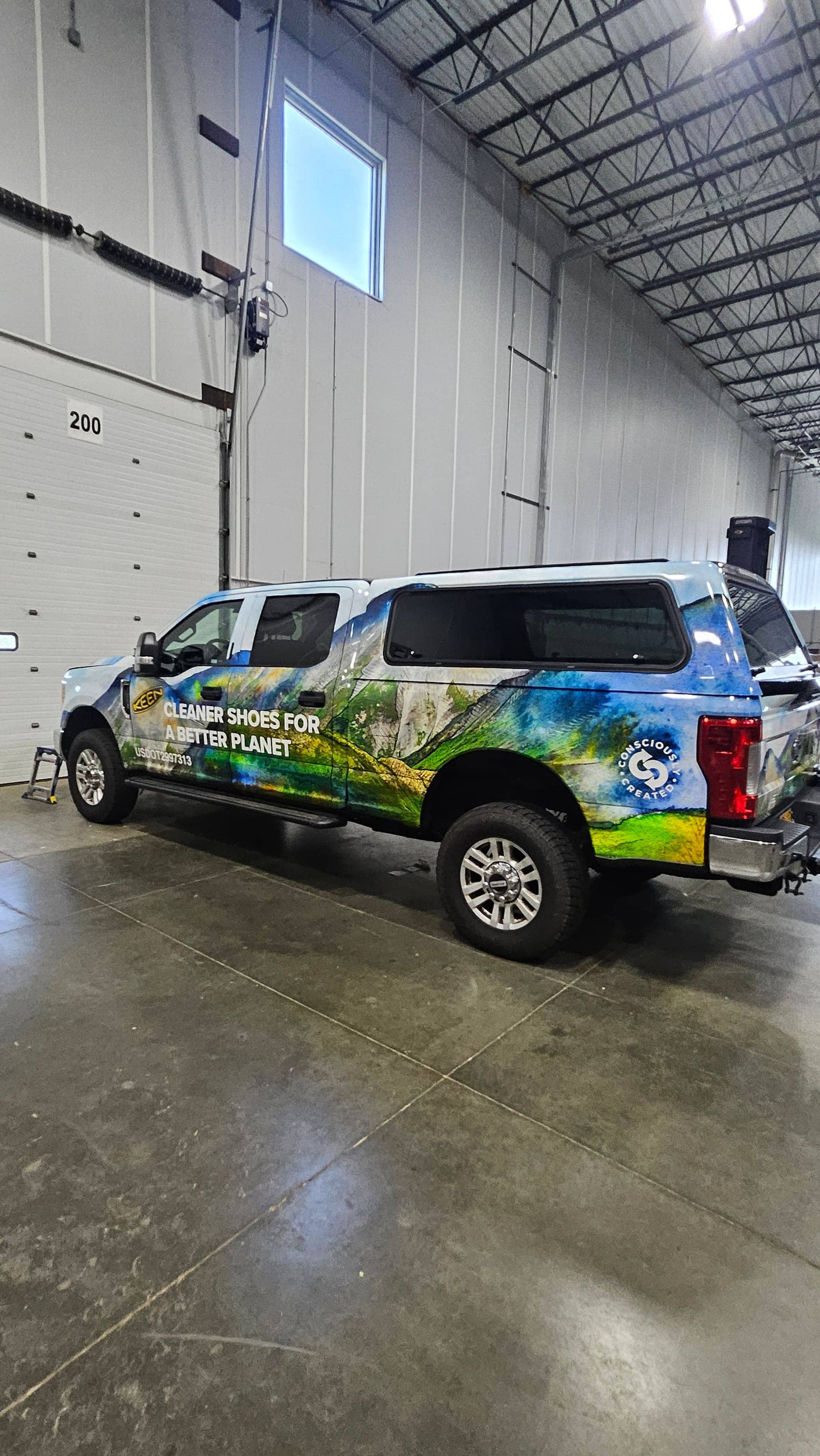 Ford F-250 and camper shell covered in a printed mountain-landscape wrap with KEEN branding, side profile