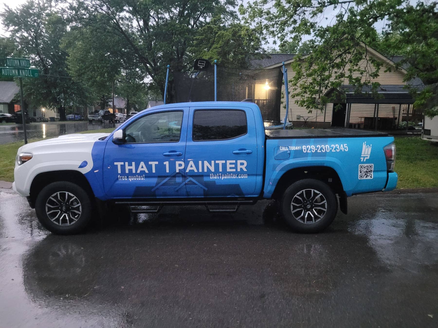 Blue Toyota Tacoma with That 1 Painter door lettering parked on a rainy street