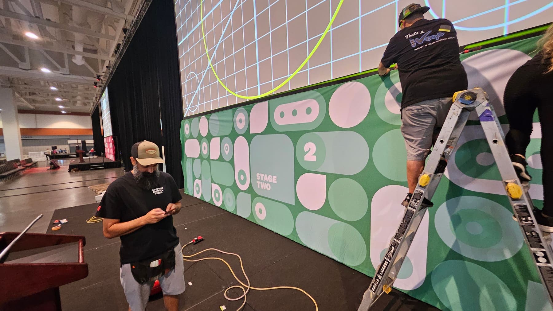 Installer on a ladder fitting a pink and green pattern banner wall at a convention stage